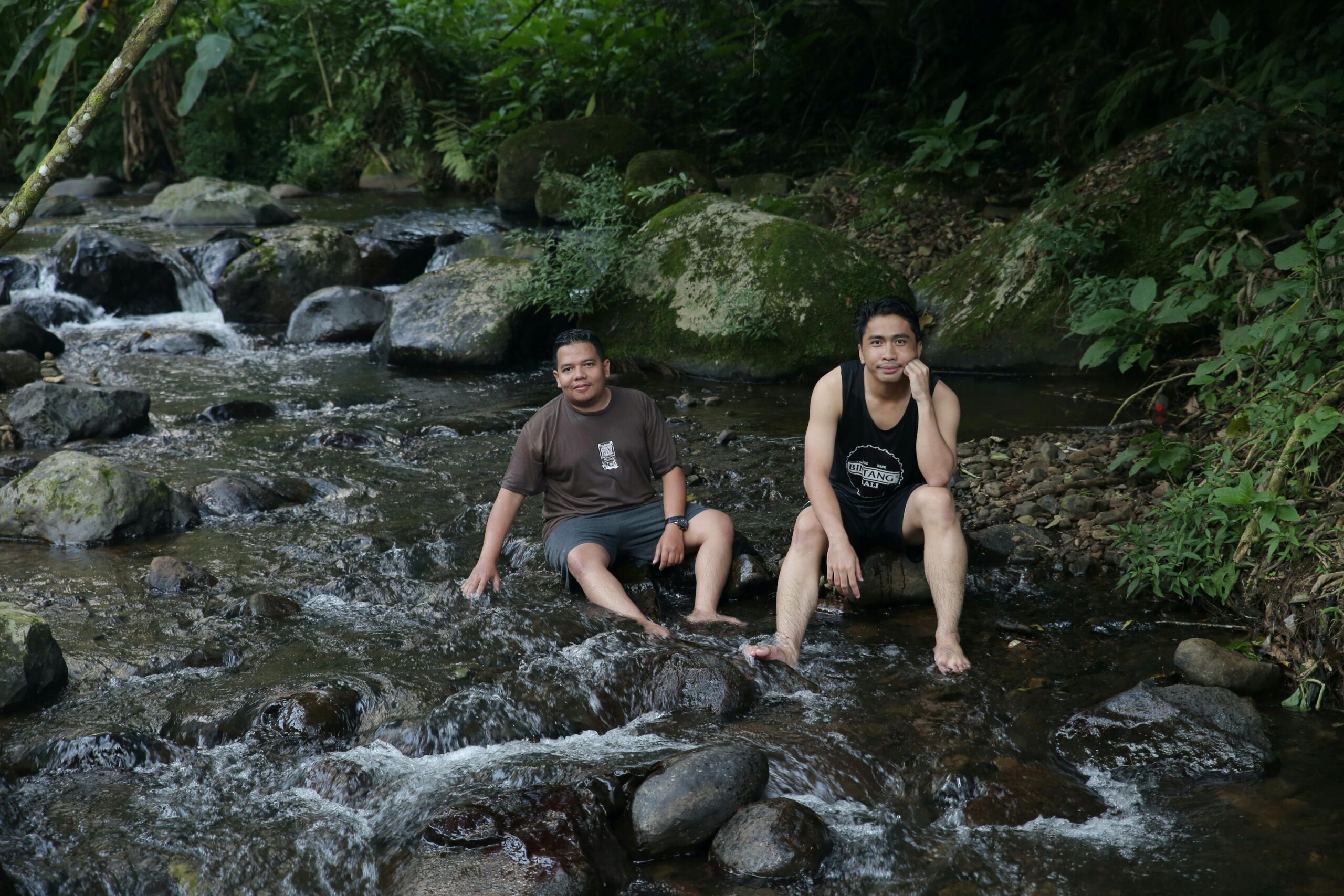 Two men enjoying a serene moment by a rocky stream surrounded by greenery.