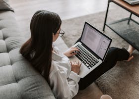 Woman working remotely with a laptop on the floor next to a sofa, enjoying comfortable home office setup.